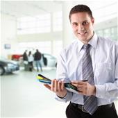 Smiling car salesman in show room with few customers in background.