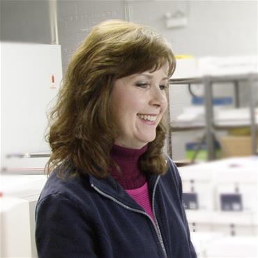 Woman (Beth) sits at her desk, working and smiling about something.